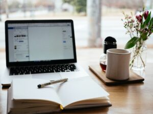 A laptop, open notebook with pen, and a coffee cup on a wooden table.