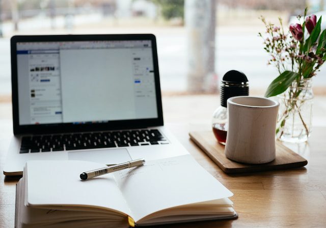 Workspace setup with laptop, notebook, coffee cup, and pen on wooden table.